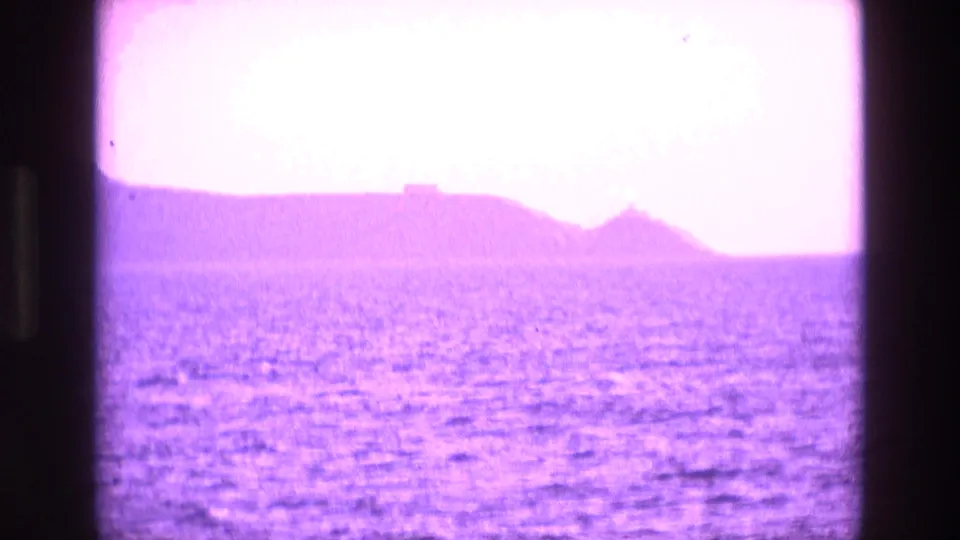 1969 Ireland coastal travel frame showing open water and a hazy shoreline, with strong exposure shift and purple color cast from 8mm film.