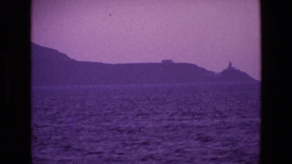 1969 Ireland home-movie coastal view across the sea toward a dark shoreline and hazy sky, framed with visible film grain and color shift.