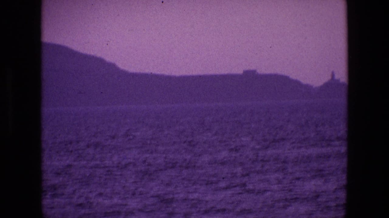 1969 8mm Ireland coastal view from the water, with rippling sea, dark hills and a small lighthouse-like form in the distance.
