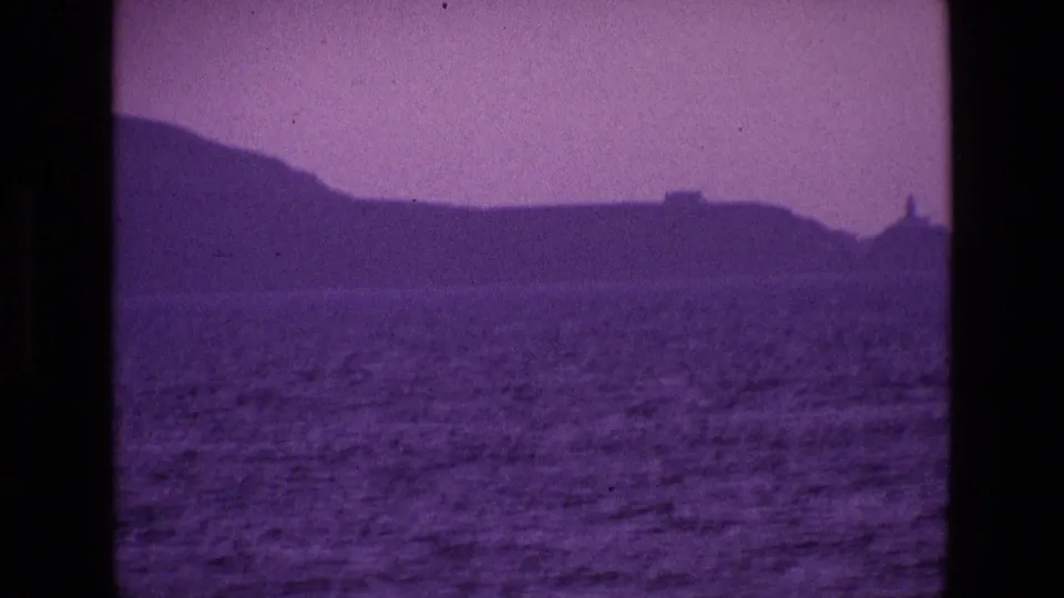 1969 8mm Ireland coastal view from the water, with rippling sea, dark hills and a small lighthouse-like form in the distance.