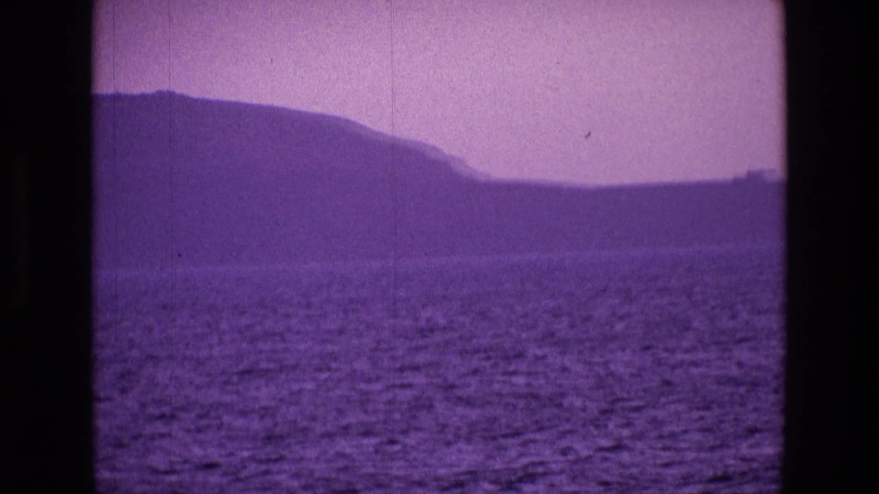 1969 8mm view of Irish coastal hills across open water, softened by purple color shift and amateur film grain.
