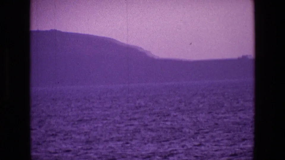 1969 8mm view of Irish coastal hills across open water, softened by purple color shift and amateur film grain.