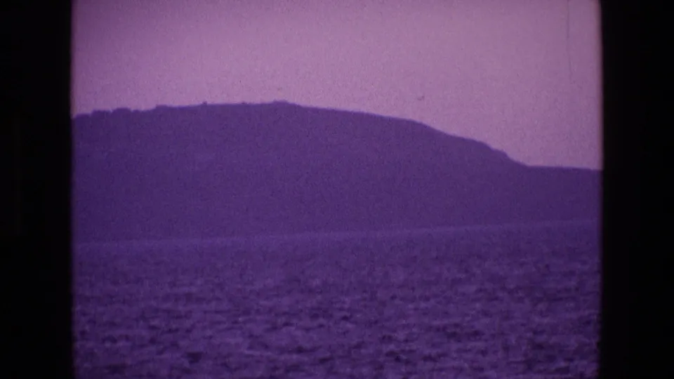 Purple-tinted 1969 home-movie frame showing an Irish headland and calm ocean from an offshore viewpoint.