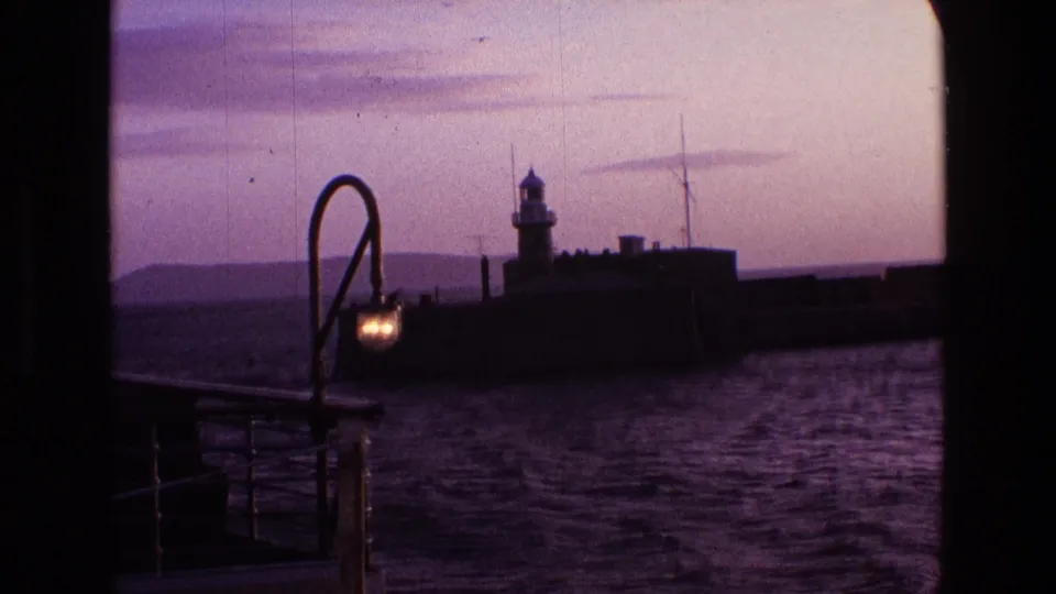 1969 Irish harbor view from a boat, with a small lighthouse on a pier and a glowing curved deck lamp in the foreground.