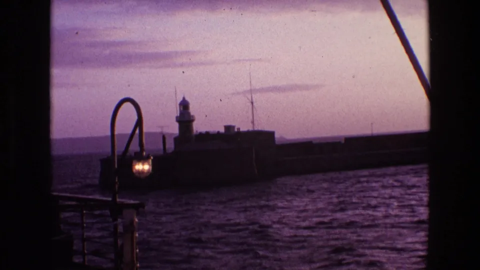 8mm frame from Ireland in 1969 showing a pier lighthouse across rippled water, seen past a lit boat lamp at dusk.