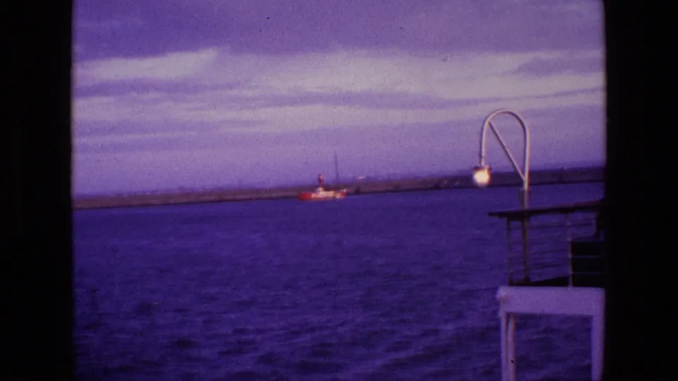 1969 Ireland 8mm frame with blue-purple sea, a red-and-white boat near the breakwater, and deck railings with a curved lamp at right.