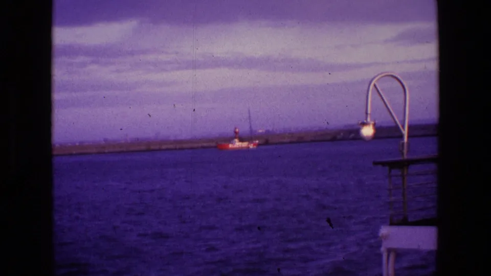 1969 Ireland 8mm frame showing a red-and-white boat on blue water, with a foreground deck lamp, railings, and clouded evening sky.