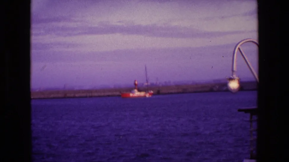 1969 Ireland 8mm frame with a small red-and-white vessel near a long breakwater, framed by a curved deck lamp and railings.