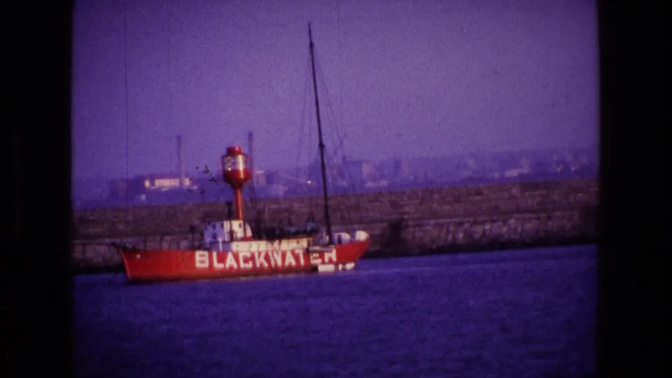 1969 Ireland 8mm frame showing the red BLACKWATER boat from the side, with mast lines and distant industrial buildings beyond the wall.