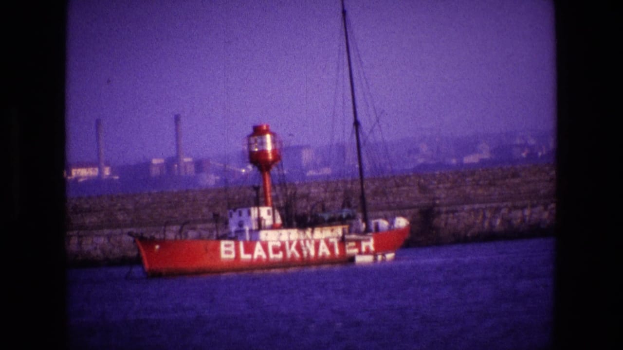 Red vessel marked BLACKWATER floating near a stone quay with industrial buildings behind it.