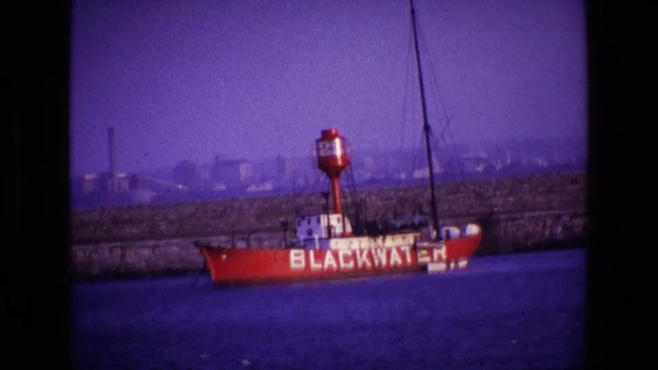 Purple-cast 8mm frame from Ireland in 1969 showing the BLACKWATER boat near a quay, with mast lines and industrial structures in the distance.