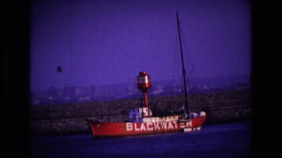 1969 Ireland harbor frame with the BLACKWATER vessel at lower right, a stone quay across the middle, and a hazy industrial background under purple-tinted film color.
