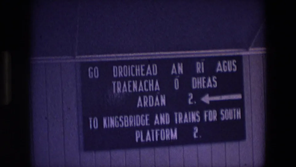 1969 Ireland 8mm frame showing a bilingual station sign directing passengers to Kingsbridge and southbound trains on Platform 2.