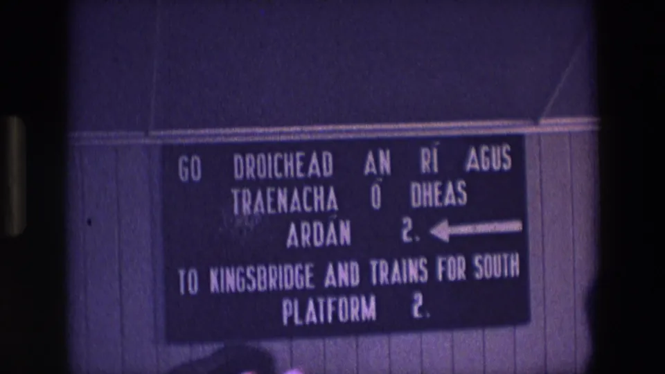 1969 Ireland home-movie frame with a purple cast, showing a wall-mounted rail sign for Kingsbridge and southbound platform access.