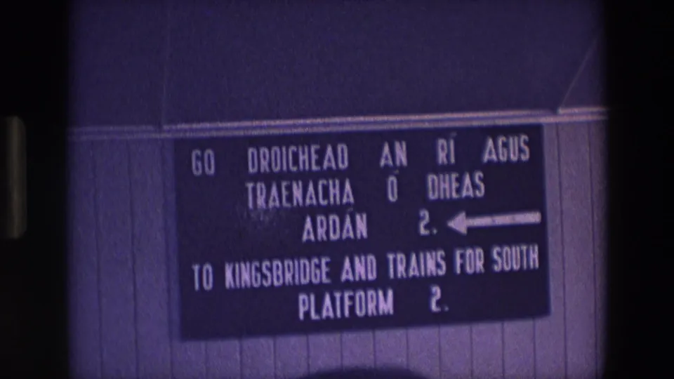 1969 Ireland 8mm frame of the bilingual Kingsbridge and southbound trains sign, with white lettering on a dark board.
