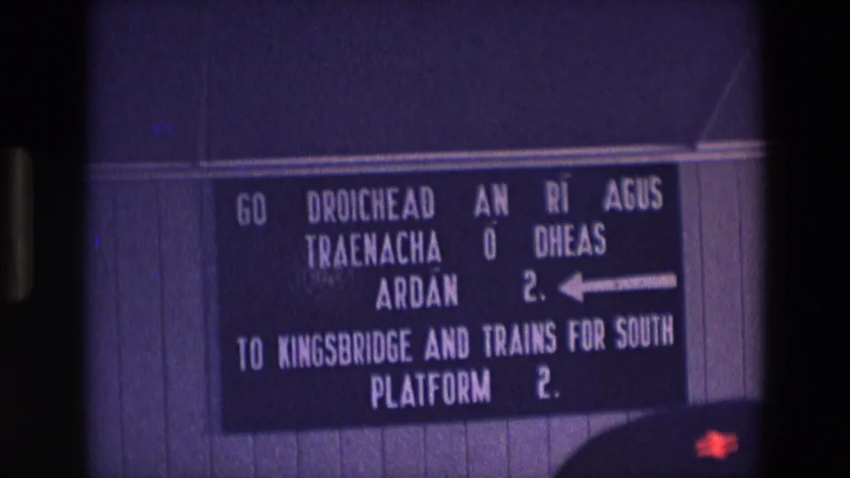 1969 Ireland 8mm frame showing a bilingual Irish and English railway station sign with a left arrow and Platform 2 directions.