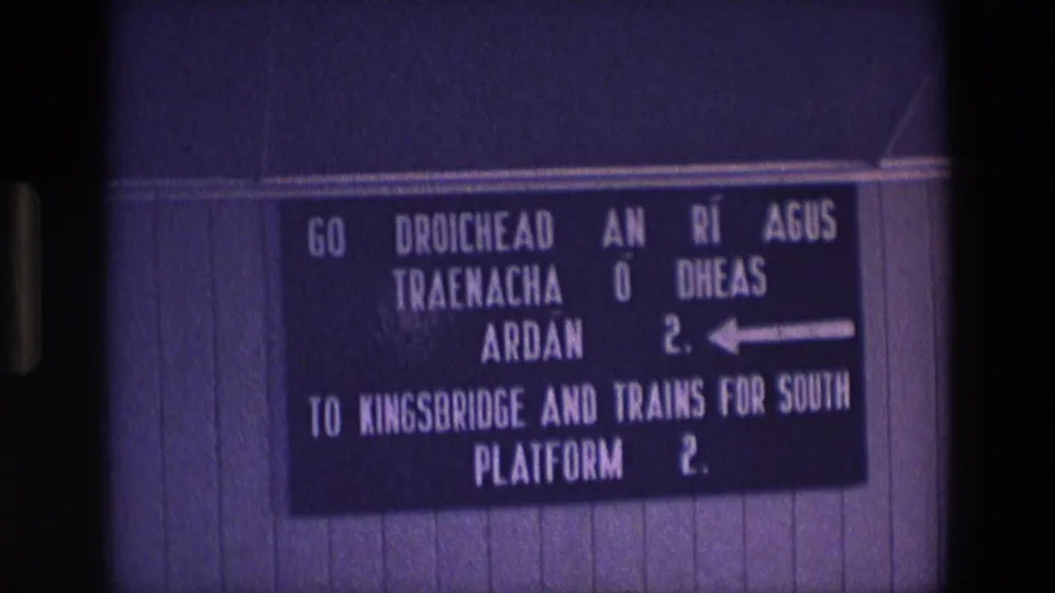 1969 Ireland 8mm still of a bilingual station direction sign for Kingsbridge and southbound trains, with soft focus and grain.