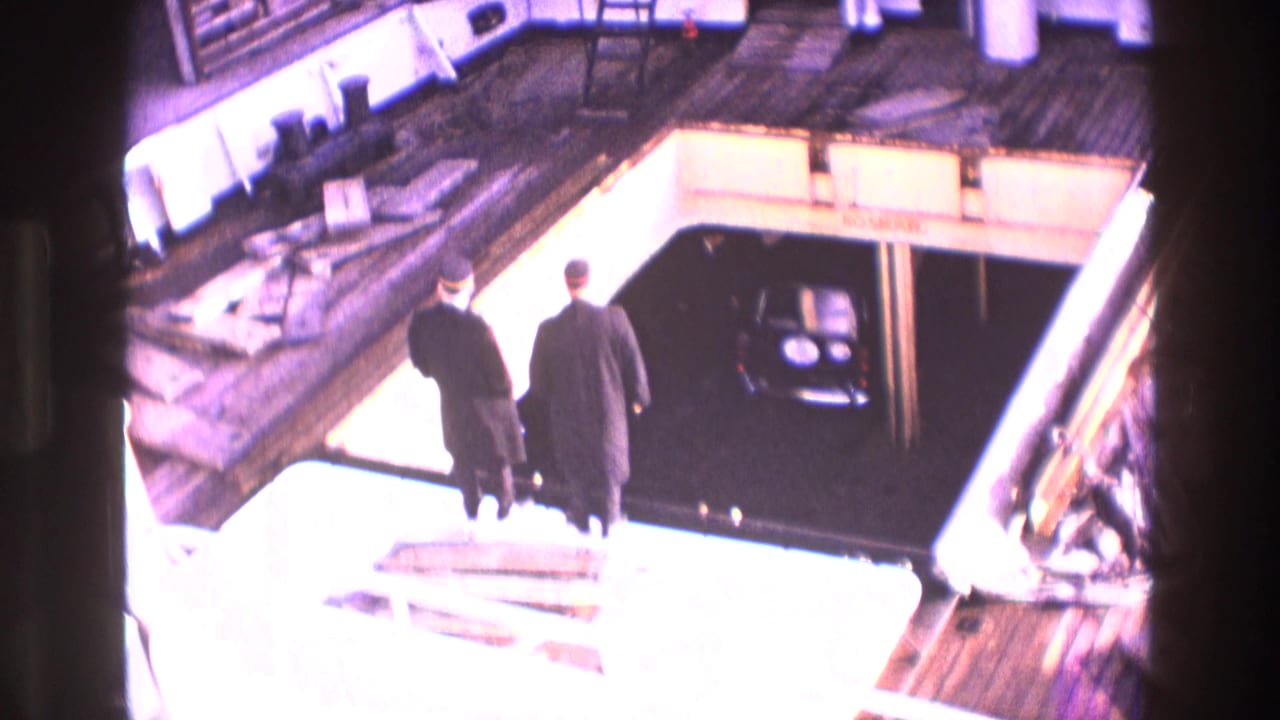 1969 Ireland 8mm frame showing two adults in dark coats crossing a ferry deck toward a lower loading bay with a black car visible.