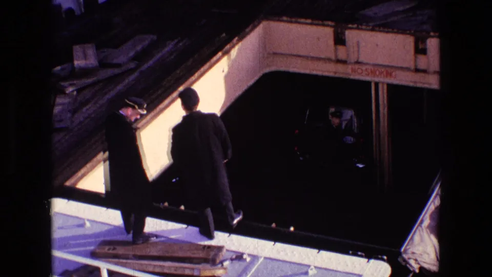 1969 Ireland 8mm frame of two adults moving along a ferry deck beside a lower car bay marked with a no-smoking notice.