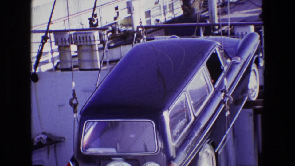Close 8mm frame of the crane-slung black vehicle beside railings, barrels, cables, and painted ship structure.