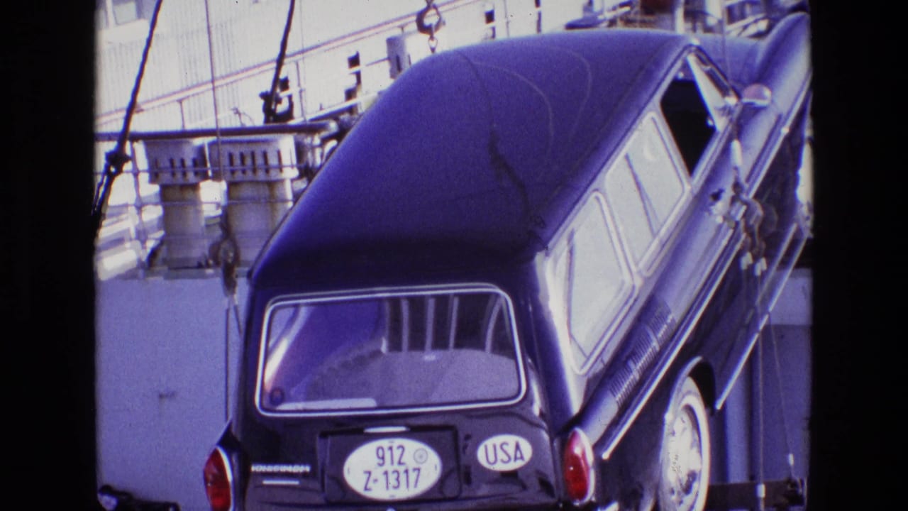 In 1969 Ireland footage, a dark station wagon with a USA plate hangs from crane chains near ship railings and dock equipment.