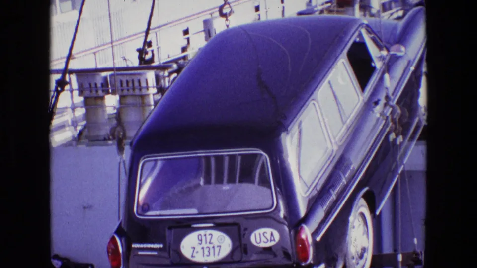 In 1969 Ireland footage, a dark station wagon with a USA plate hangs from crane chains near ship railings and dock equipment.