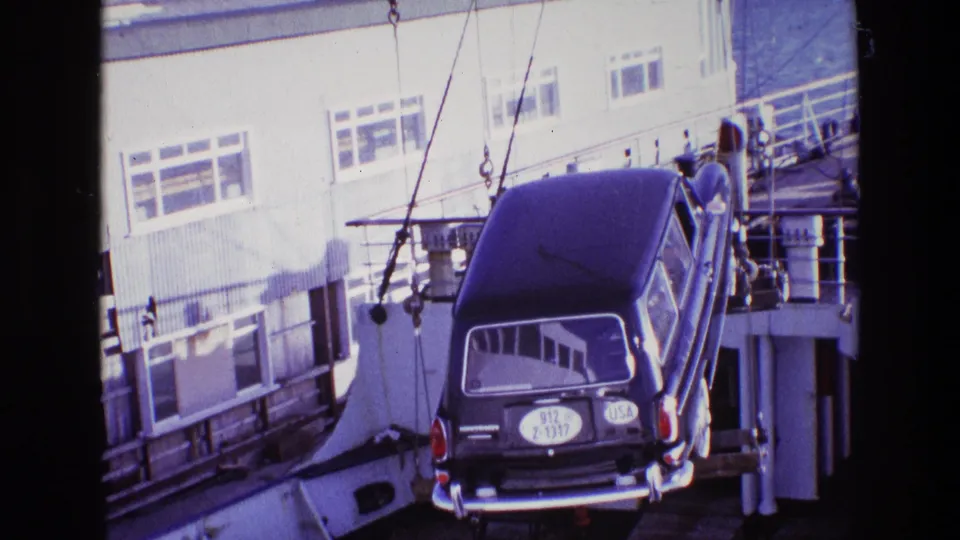 The crane lowers the dark station wagon near a dockside building, with windows, railings, and harbor equipment in the background.