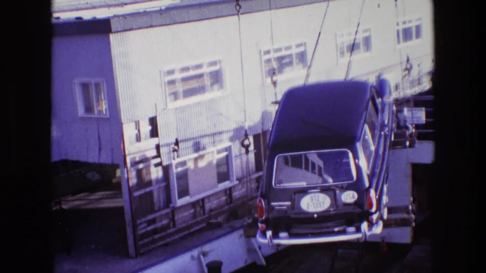 The station wagon is nearly level with the dock area, still held by crane chains against a backdrop of port buildings and railings.