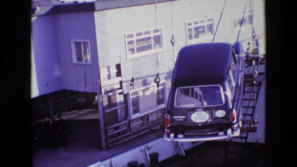 1969 Ireland dockside view of a black hearse hanging from hoist cables near a corrugated building as men watch from below.
