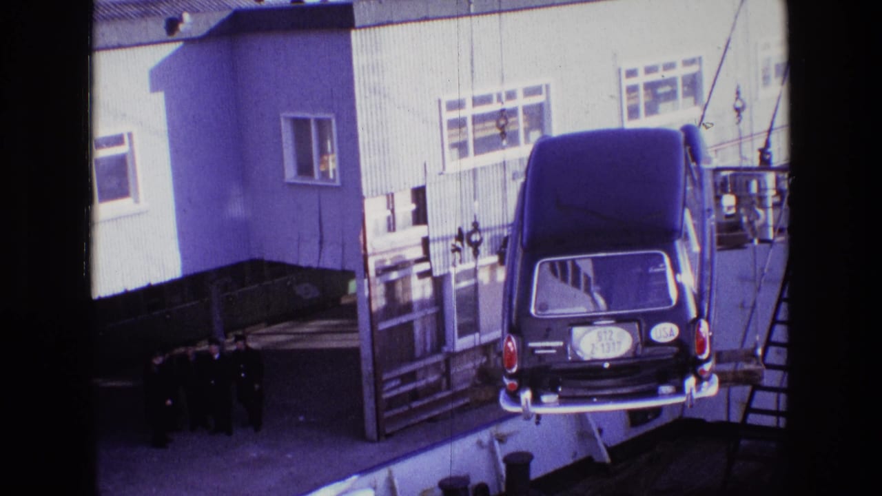 Rear of a suspended black hearse near rigging, a ladder, and a white building at a dock.