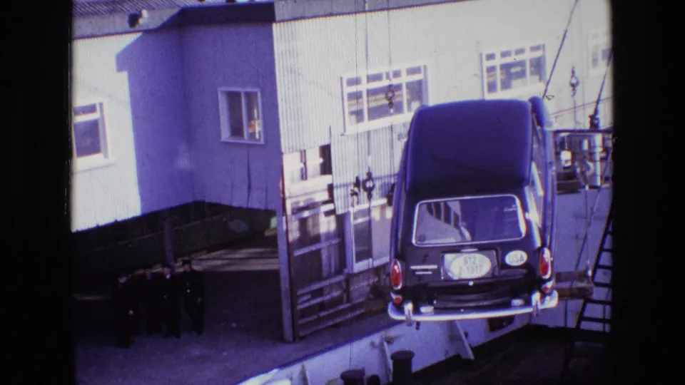 The hearse remains suspended near ship or dock rigging, with a white industrial building and a small group of men visible below.