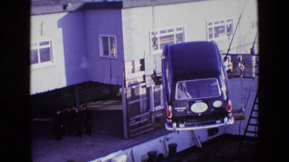 A black hearse is angled in midair beside the dock, with rigging lines and a group of men visible against the industrial building.