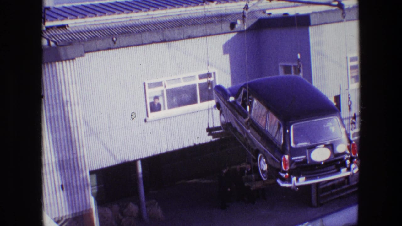 In Ireland in 1969, a black hearse hangs from crane rigging outside a corrugated industrial building as a man watches from a window.