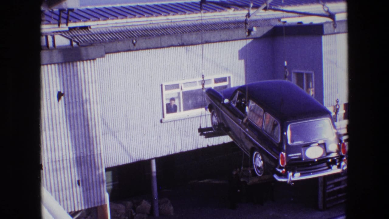 Crane holds a black hearse above a platform outside a metal-sided building.