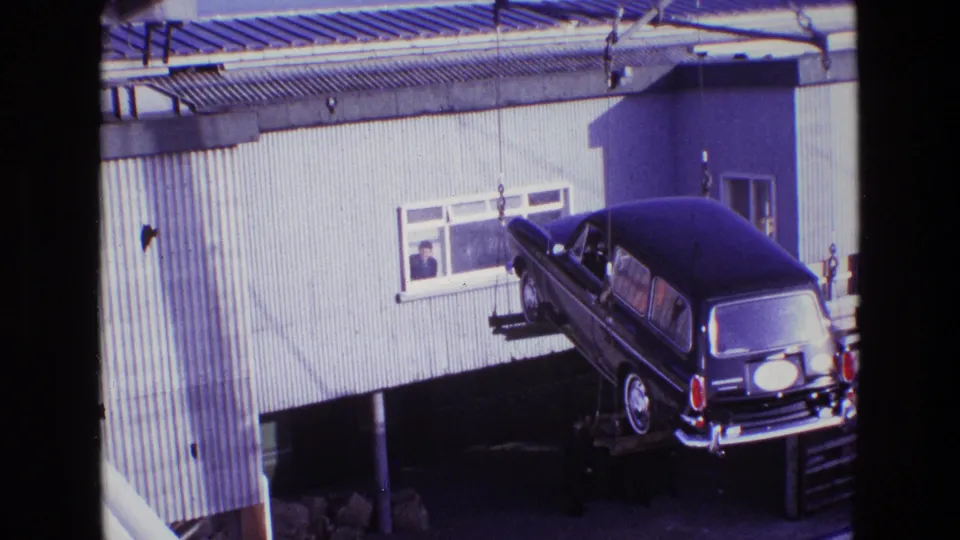 The suspended black hearse is angled beside the industrial wall, with crane cables above and an observer visible through a window.