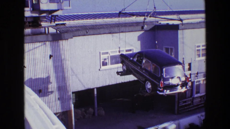 A black hearse is lowered or shifted by crane outside the building, with workers below and a window observer in the background.