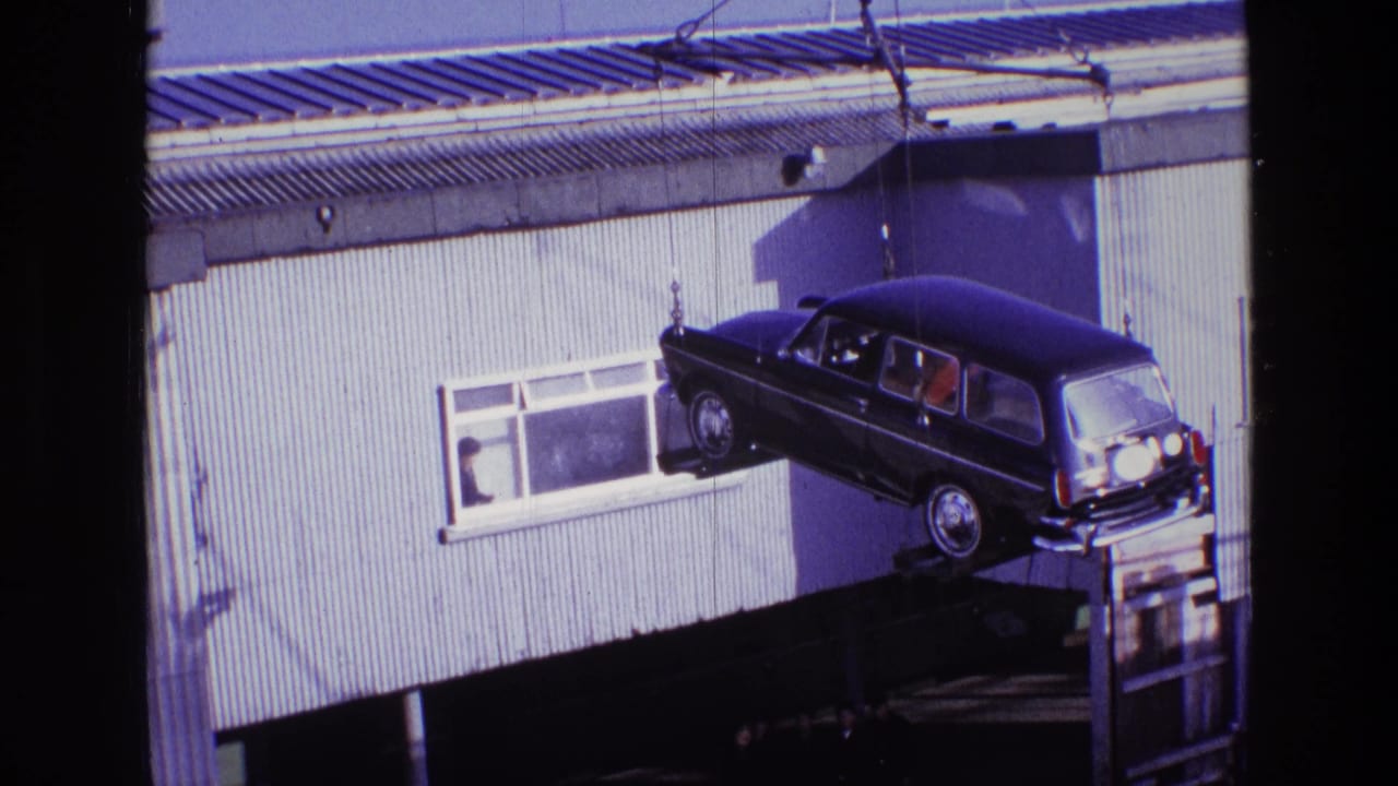 The dark station wagon remains suspended by taut crane lines against a corrugated building in Ireland in 1969.