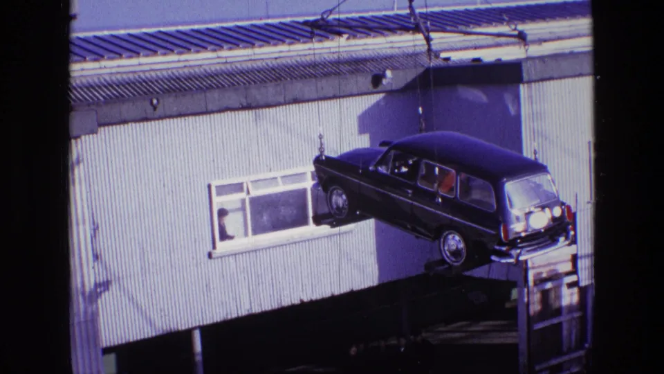 The dark station wagon remains suspended by taut crane lines against a corrugated building in Ireland in 1969.