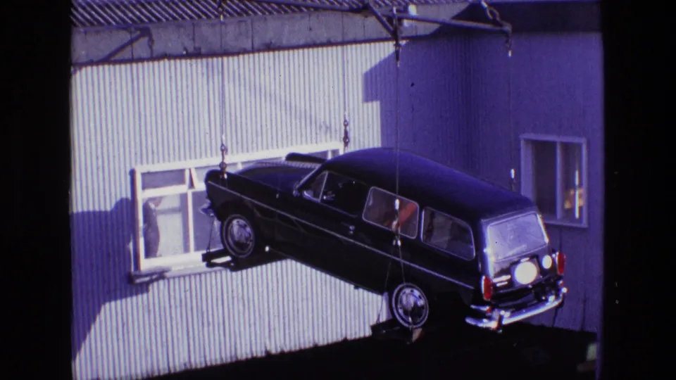 At the end of the clip, the dark estate car hangs level from crane cables near a corrugated wall and small window.