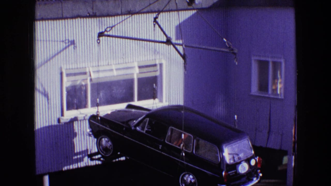 In Ireland, 1969, a black classic station wagon hangs from crane cables near a corrugated dock building and windows.