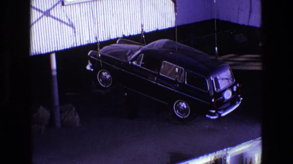 The black station wagon remains suspended from crane lines at an Irish dock, with strong shadows and metal siding behind it.