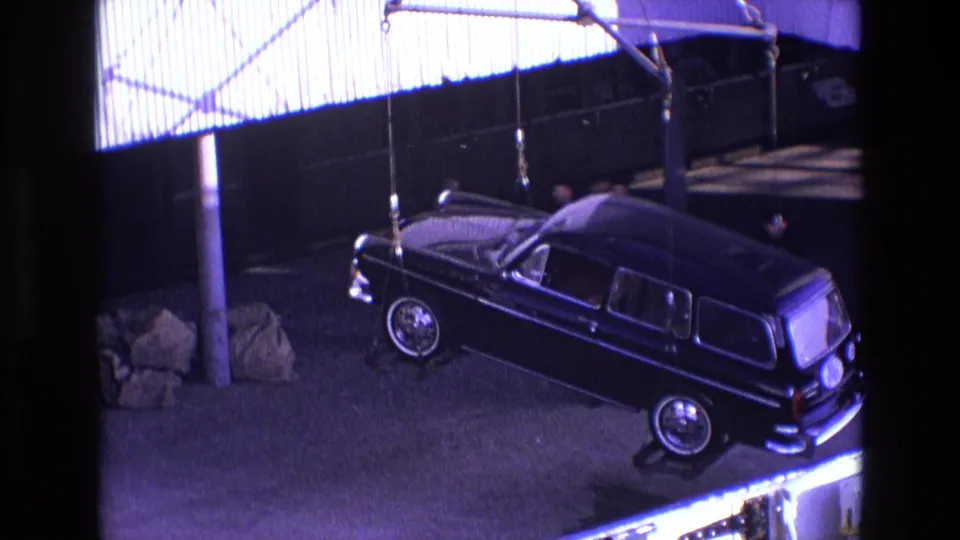 A crane holds the black classic station wagon above the dock, with chrome trim, wheels, and a nearby pole visible in the frame.