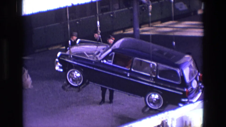 Men in dark clothing walk near the suspended black station wagon as it hangs from crane cables at an Ireland dock in 1969.