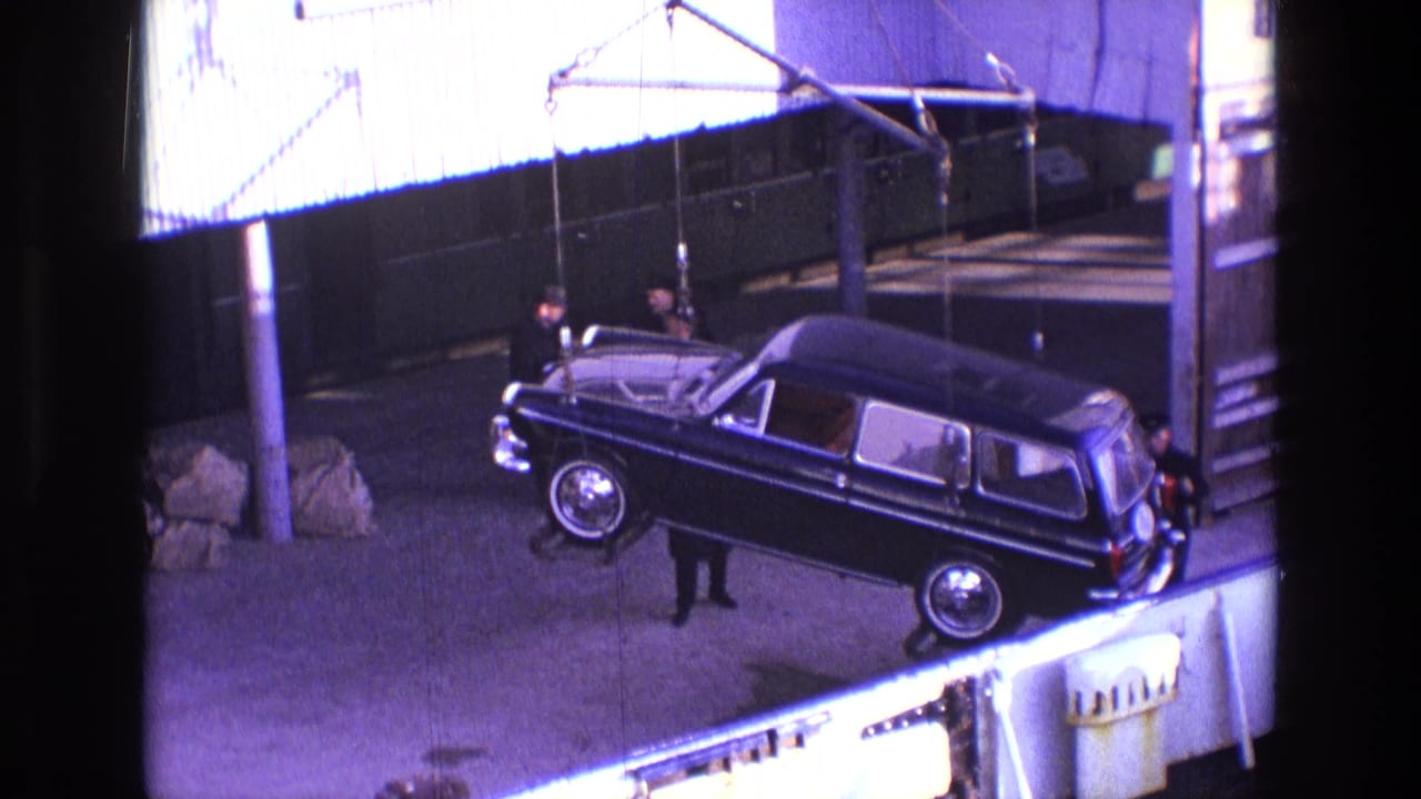 In 1969 Ireland footage, a black hearse-like vehicle hangs from crane cables while men stand beside it in an industrial yard.