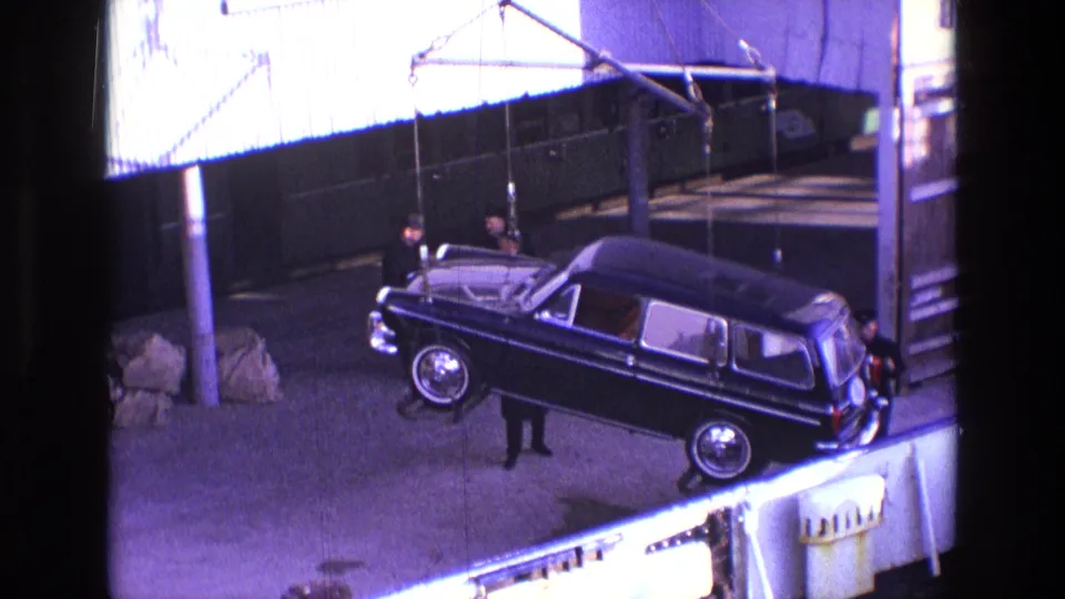 In 1969 Ireland footage, a black hearse-like vehicle hangs from crane cables while men stand beside it in an industrial yard.