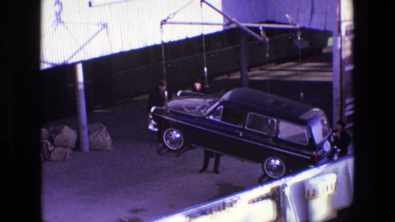 The black hearse-like vehicle remains suspended from cables, with one man below and industrial cladding behind it in the 1969 Ireland clip.