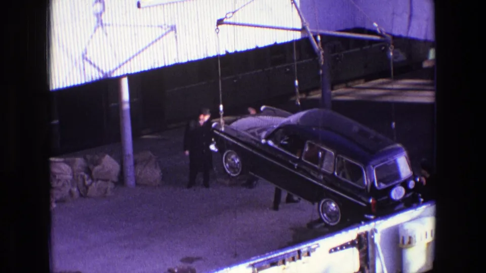 Several men in dark coats guide or watch the suspended black vehicle as it shifts position in an Irish industrial yard in 1969.