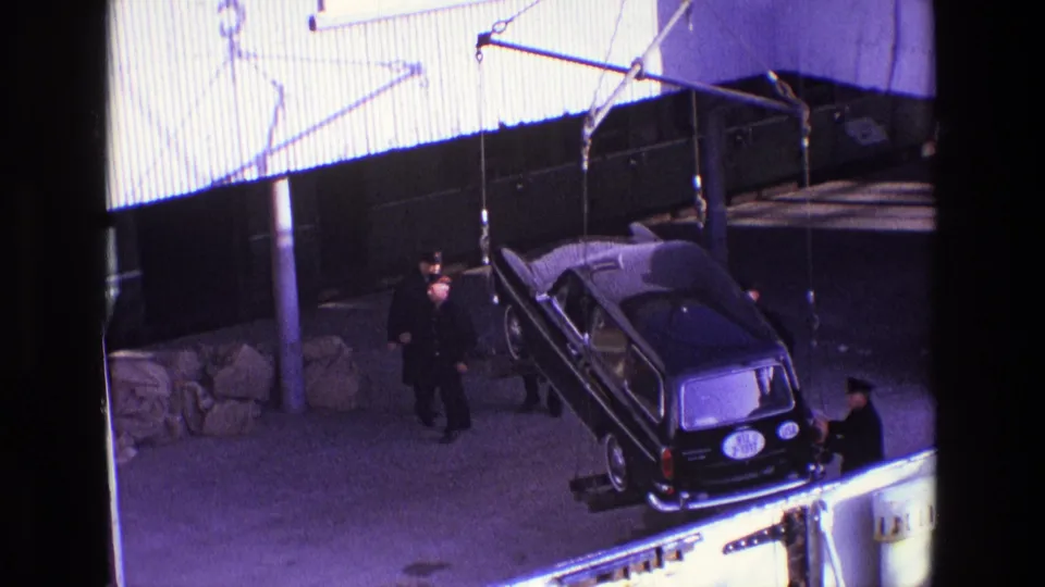 The rear of the black hearse-like vehicle faces the camera as workers stand nearby during the crane lift in 1969 Ireland footage.