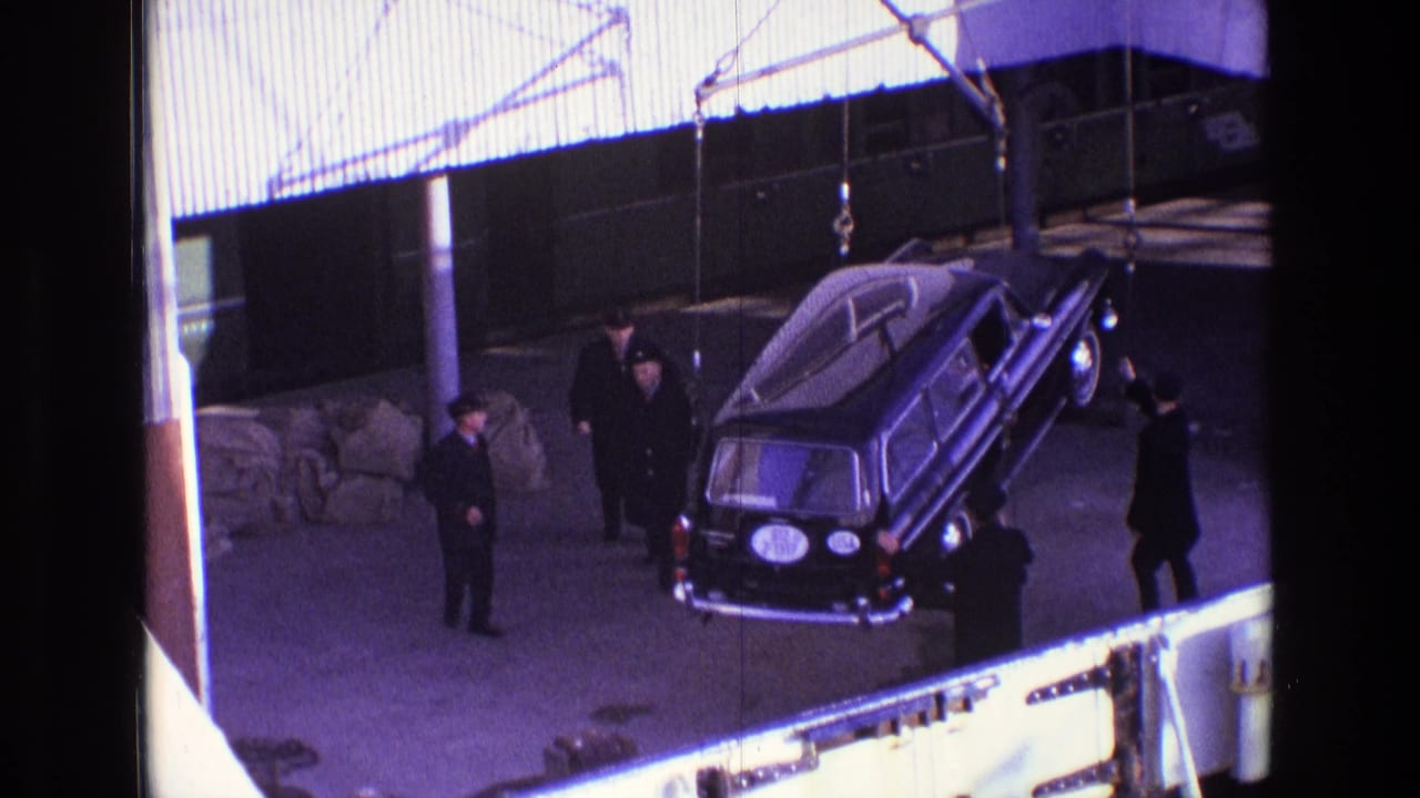 Hearse tilted under crane rigging beside men on a dock.