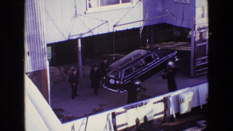 A black hearse is raised higher by crane at a dockside industrial setting, with men positioned around the vehicle.